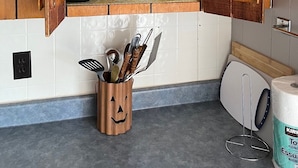 Una cocina con armarios de madera, un estante con cristalería y platos, y un portalapapeles con diseño de calabaza de Halloween.