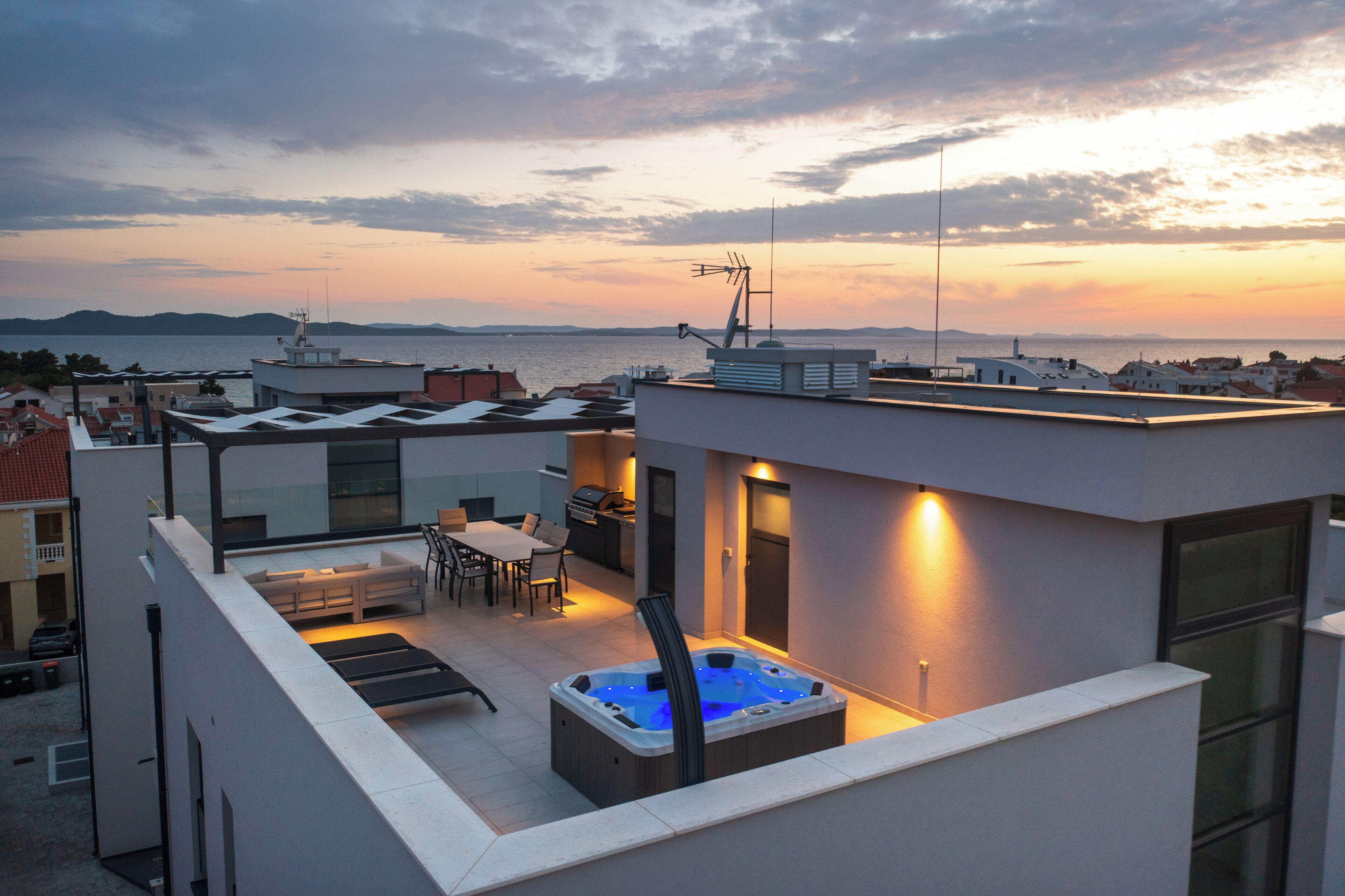 En tagterrasse med jacuzzi, udendørs møbler og havudsigt i solnedgangen.