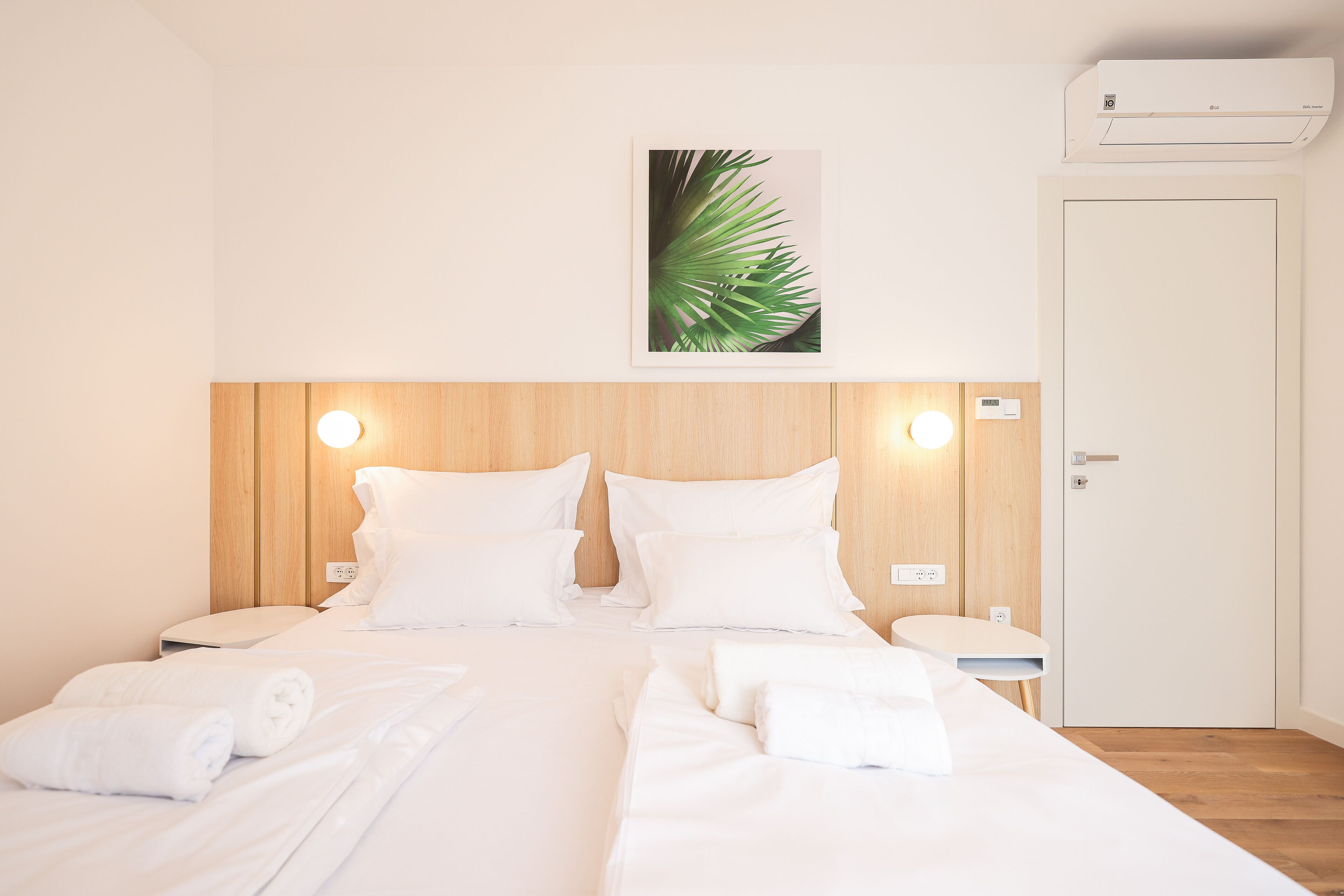 A neatly made bed with white linens and towels, a wooden headboard, a wall-mounted air conditioner, and a small bedside table with a lamp.
