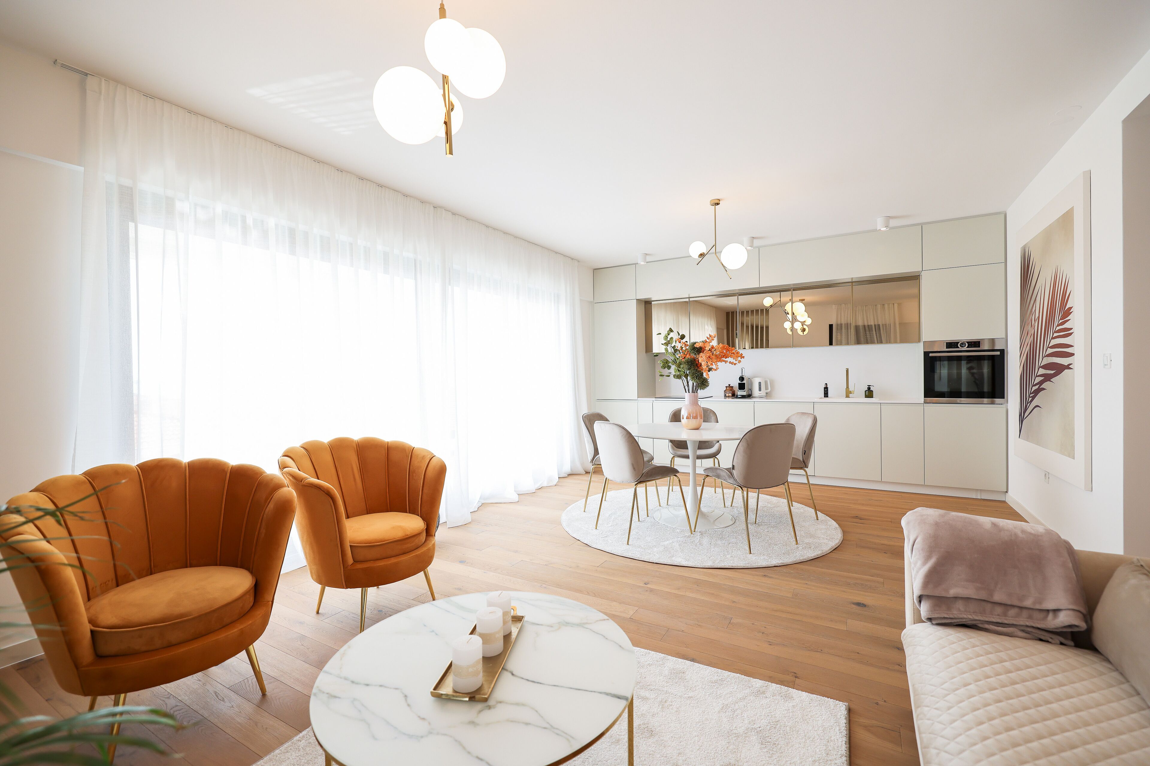 A modern living room with a marble coffee table, two orange armchairs, a beige sofa, and a dining area with a white table and chairs.