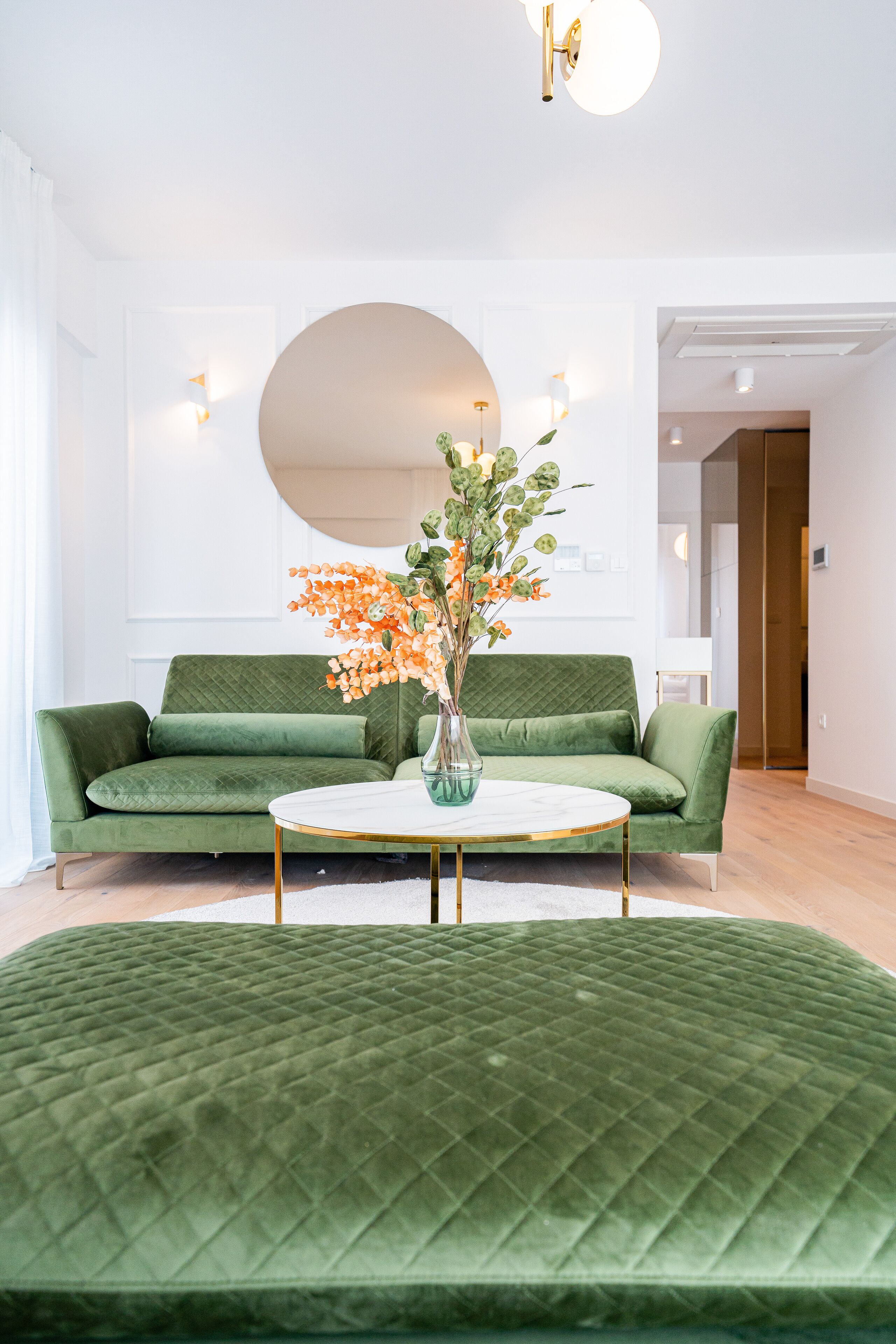 Un sofá verde con una mesita redonda al frente, un espejo grande en la pared y un jarrón con flores sobre la mesa.