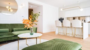 A modern living room with a green sofa, a round coffee table, and a green ottoman. There is a kitchen area with white cabinets and a bar counter. - Adria Concept boutique apartments (Zadar)