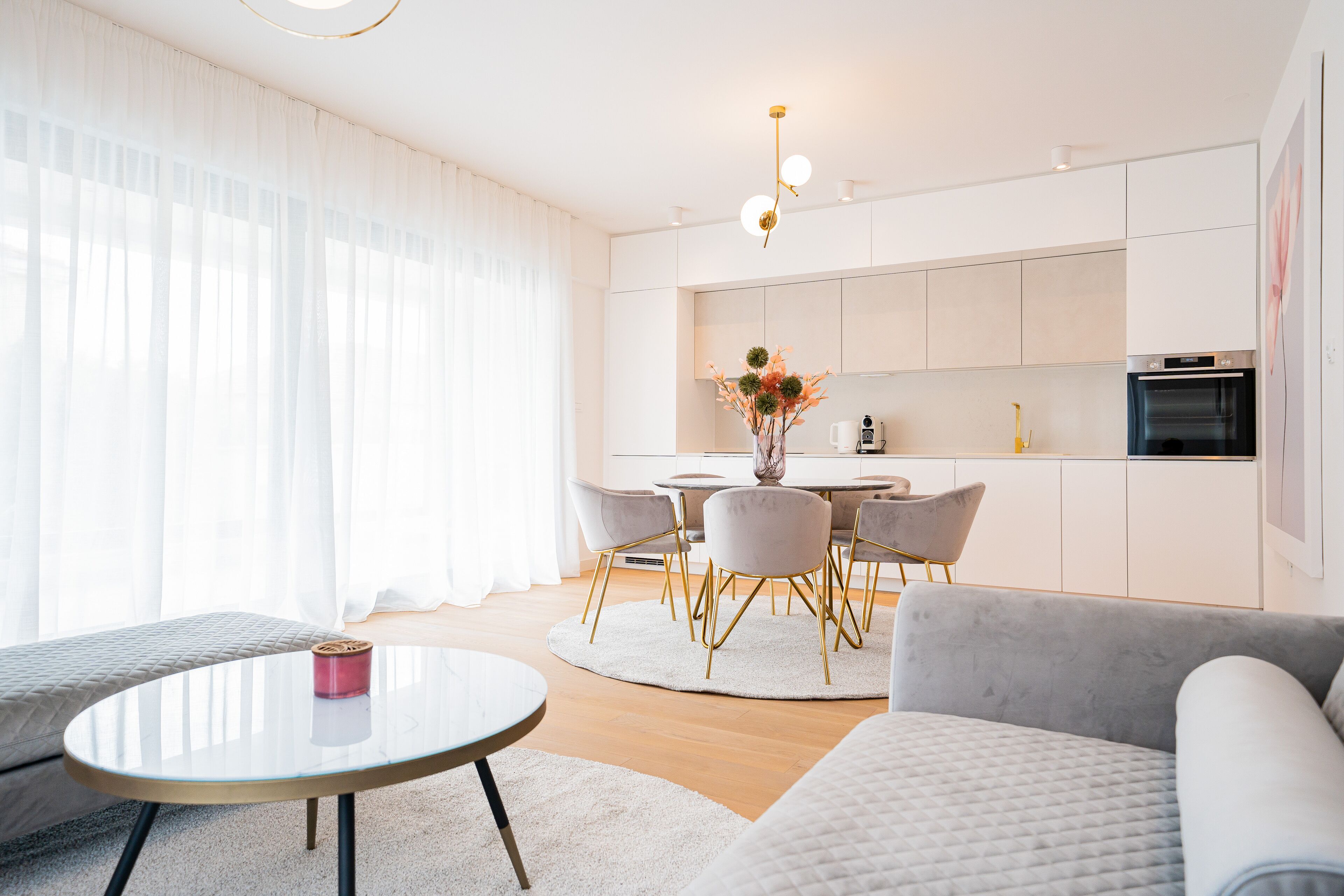 A modern living room with a round glass table, a grey sofa, and a dining area with a white table and chairs.