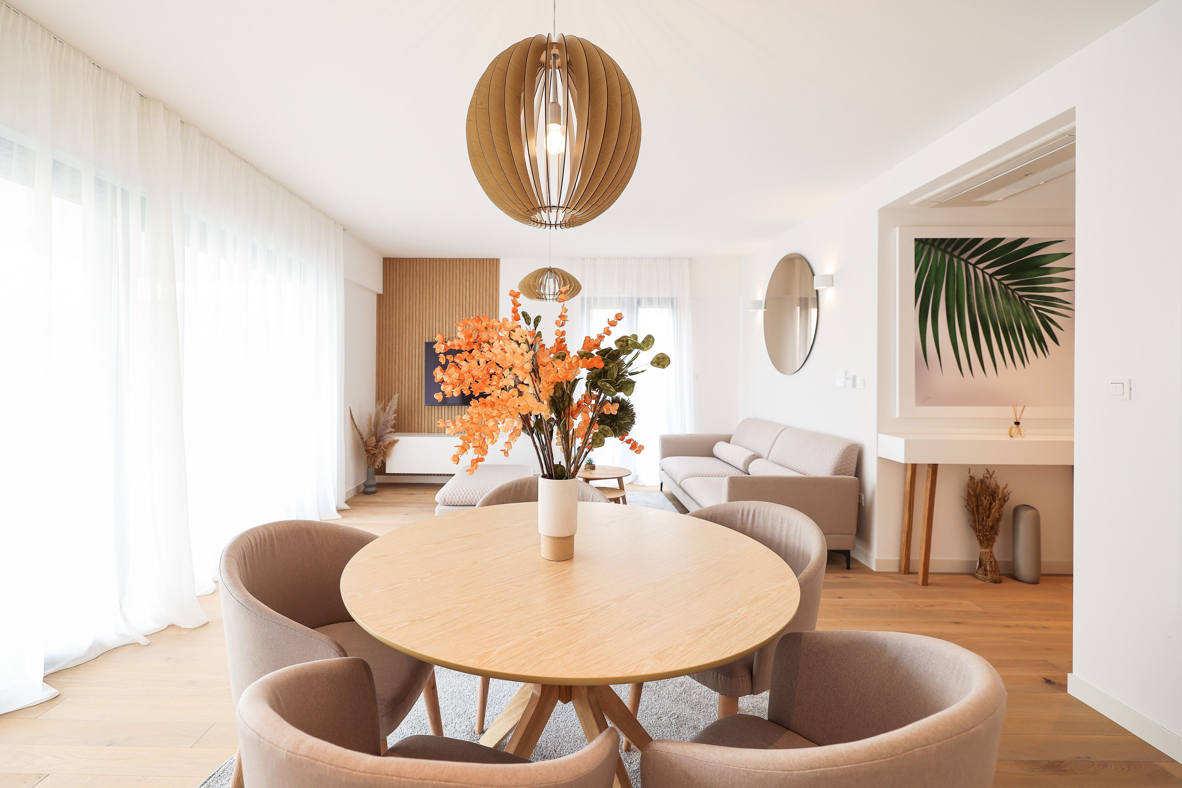 A modern living room with a round wooden table, beige chairs, and a decorative vase with orange flowers.