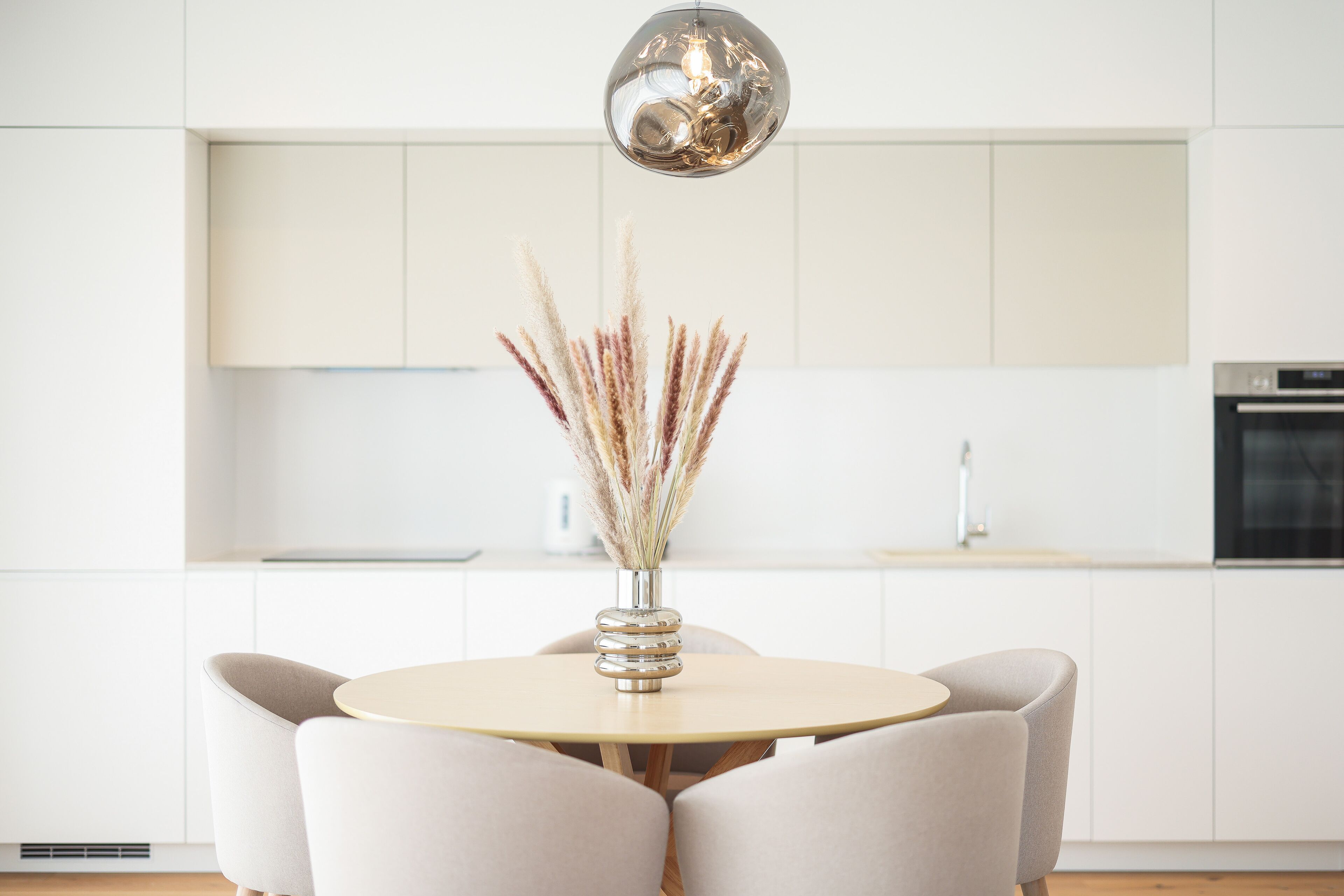A modern kitchen with white cabinetry, a round table with chairs, and a decorative vase with dried plants.