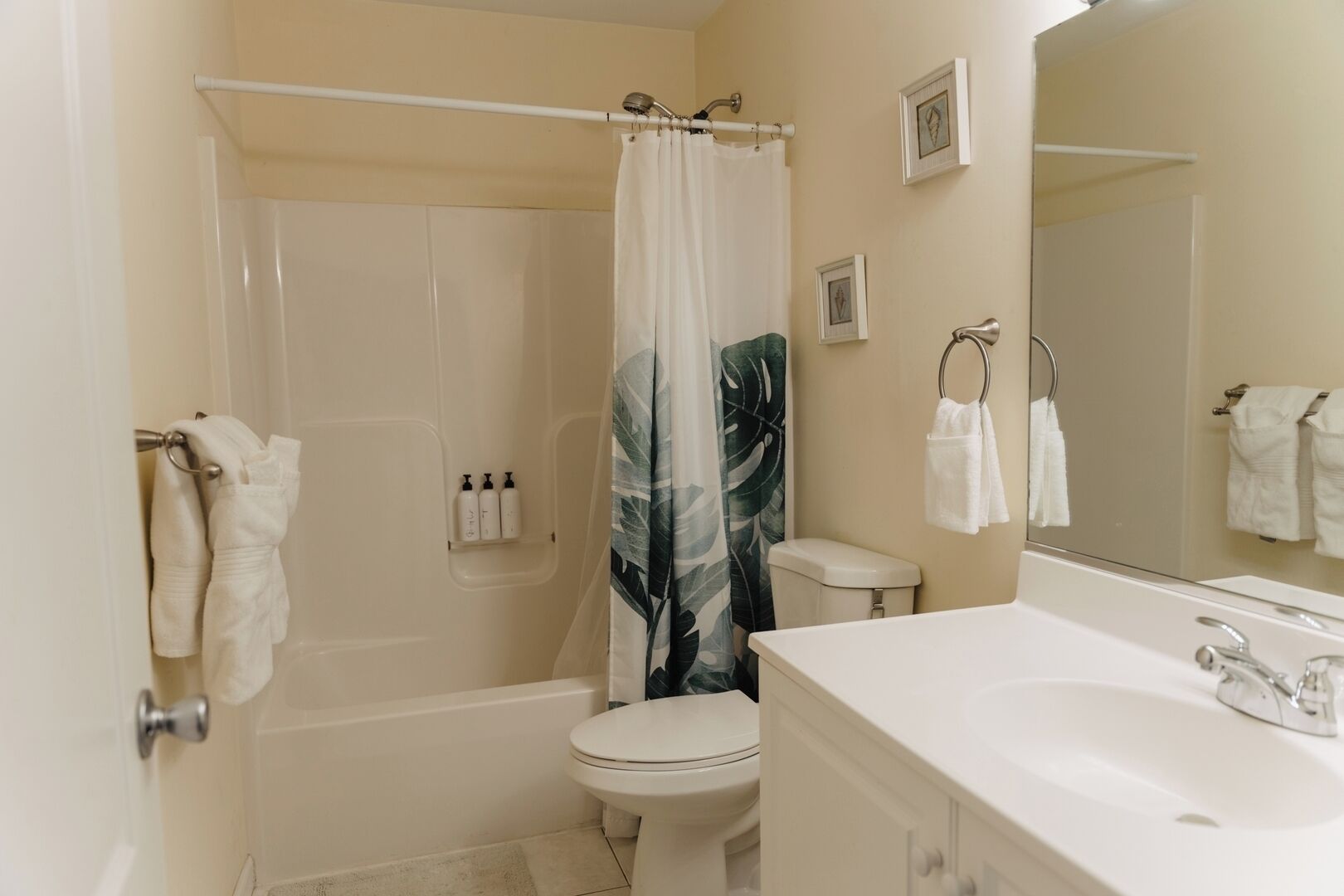 Une salle de bain avec une baignoire, des toilettes et un lavabo. Des serviettes sont suspendues sur un porte-serviettes, et il y a un rideau de douche orné d’un motif de feuilles.
