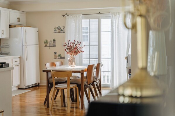 Una cocina moderna con armarios blancos, un refrigerador blanco, una mesa de comedor con sillas y una ventana con cortinas translúcidas.