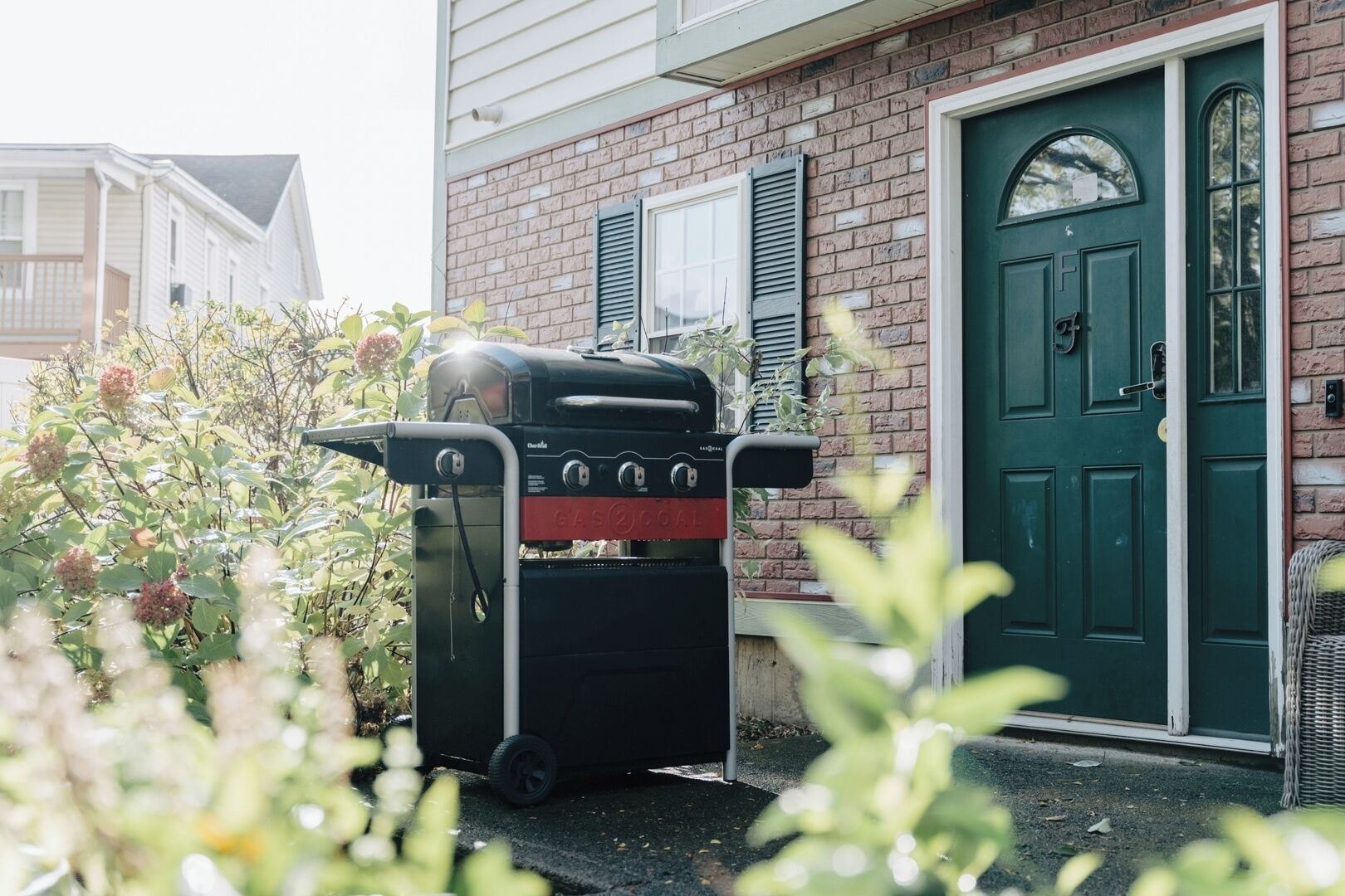 Un barbecue noir, avec une bouteille de gaz rouge, est placé devant une maison en briques dotée d’une porte verte et de volets blancs.