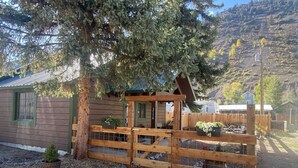Un chalet rustique avec une porte en bois, un petit espace clos entouré de plantes en pot et un paysage montagneux en arrière-plan.