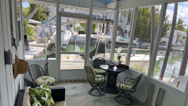 A covered patio with a view of a marina, featuring a sofa, chairs, a small table, and a glass door leading to the outside.