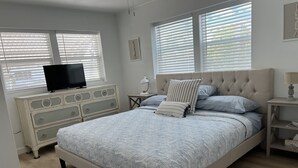 A bedroom with a large bed, a dresser, a TV, and two windows with blinds.