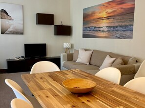 Ein modernes Wohnzimmer mit einem Holzesstisch, weißen Stühlen, einem beigen Sofa, einem Flachbildfernseher und zwei gerahmten Bildern an der Wand.