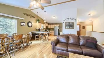 A spacious living area with a brown leather sofa, wooden dining table, and chairs. A ceiling fan and recessed lighting are visible.