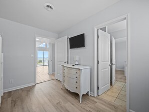 Una habitación con piso de madera, paredes blancas, un tocador blanco, un televisor de pantalla plana y una puerta que da a un balcón.