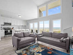 A spacious living area with a kitchen, grey sofas, a colorful rug, and large windows offering a view of the sea.