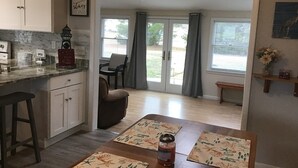 A kitchen with a marble countertop, a sign that says "OPEN", and a ceiling fan. There is a dining area with a table and chairs, and a bench.