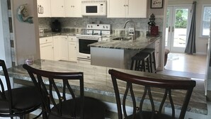 A modern kitchen with white cabinets, a built-in microwave, a double oven, and a granite countertop. There are two bar stools and a dining table.