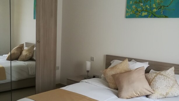A bedroom with a large bed, a bedside table, a lamp, a painting, and a mirror. - Modern 2 Bedroom Suite with Kitchen, A/C and Ample Parking (Naxxar)