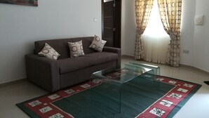 A living room with a brown sofa, patterned cushions, a glass coffee table, a red and green patterned rug, a painting on the wall, and a window with patterned curtains. - Modern 2 Bedroom Suite with Kitchen, A/C and Ample Parking (Naxxar)