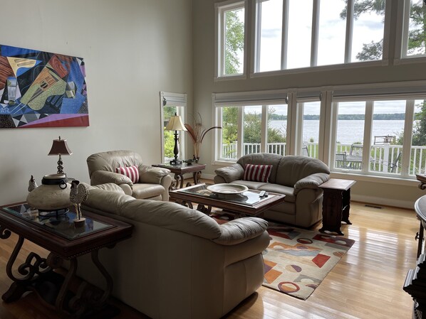 A spacious living room with a large window overlooking a lake, furnished with leather sofas, a coffee table, and a painting on the wall. - Cozy, Private Water Front Home with Long Pier (Lancaster)
