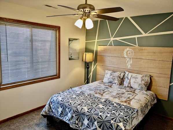 Ein Schlafzimmer mit einem Bett, einem Deckenventilator, einem Fenster mit Jalousien und einer Wand mit geometrischen Mustern.