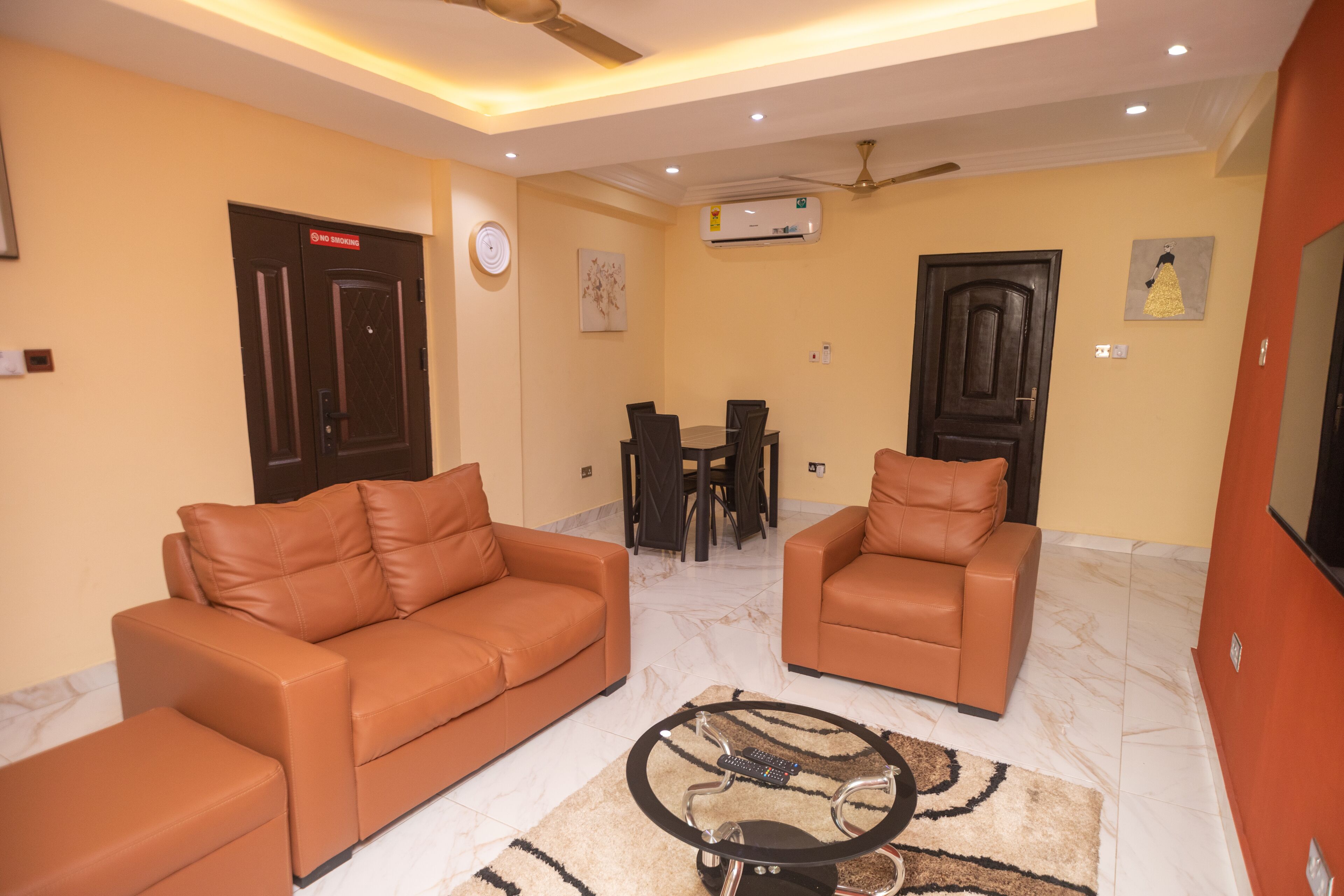 A living room with two brown leather sofas, a glass coffee table, a dining table with chairs, and an air conditioning unit on the wall.