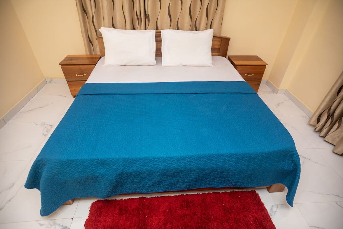 A bed with a blue cover, two white pillows, wooden nightstands, and a red rug.