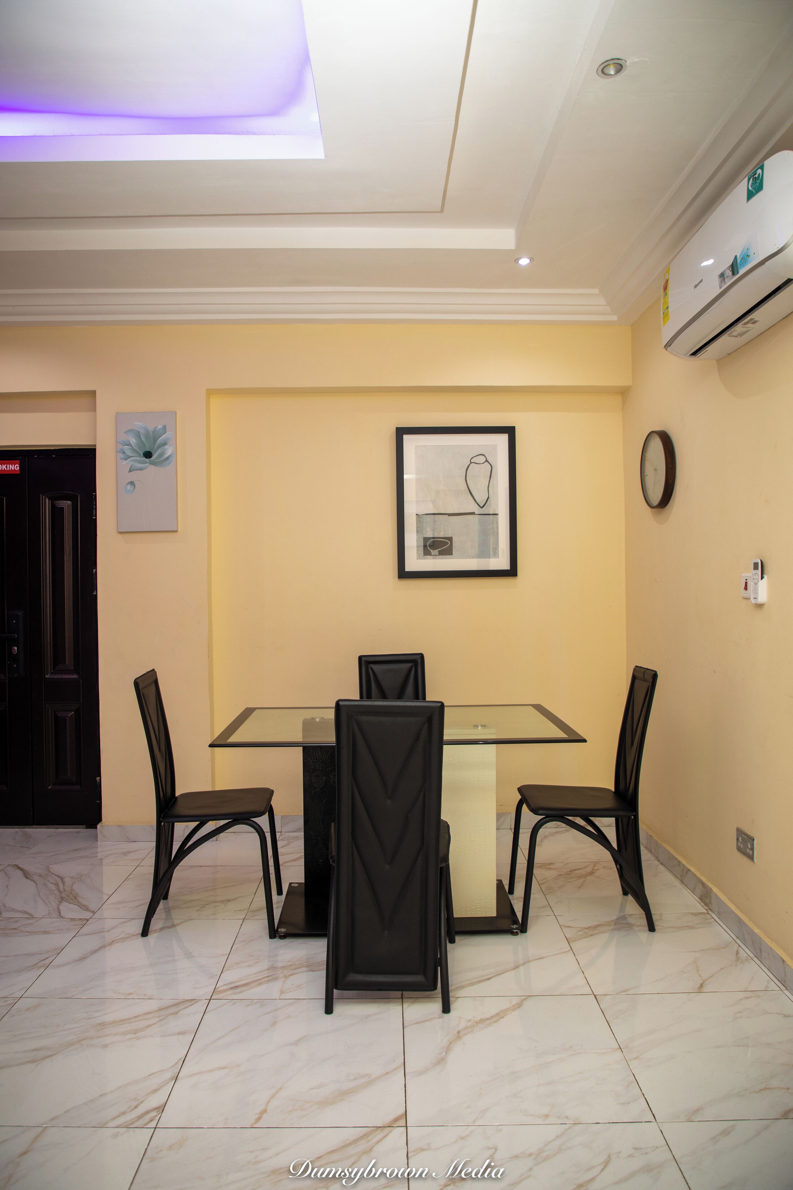 A dining area with a glass table, four chairs, a framed artwork, an air conditioning unit, and a clock on the wall.