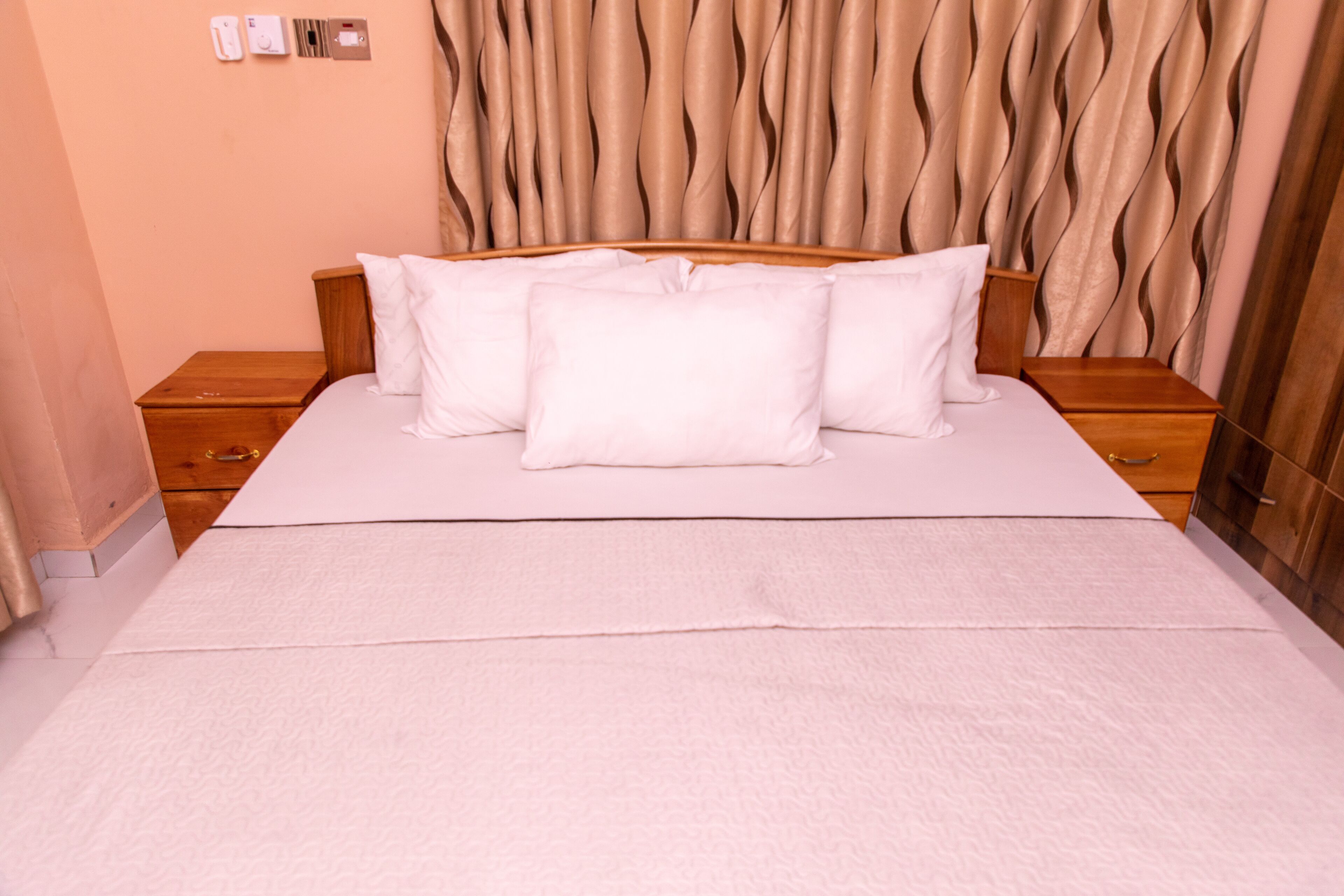 A neatly made bed with white linens and pillows, a wooden nightstand with a drawer, and a wall-mounted light switch.