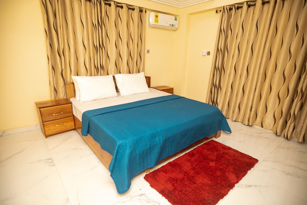 A bedroom with a bed, a nightstand, a wooden drawer, a red rug, and beige curtains.