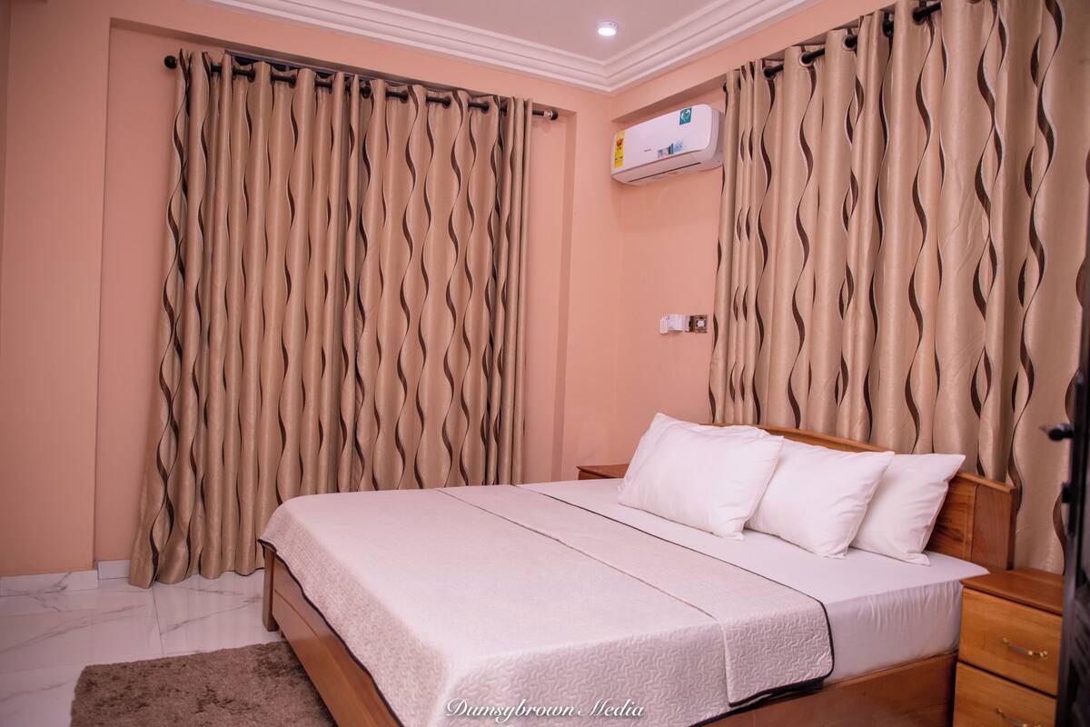 A bedroom with a bed, curtains, a nightstand, and a wall-mounted air conditioner.