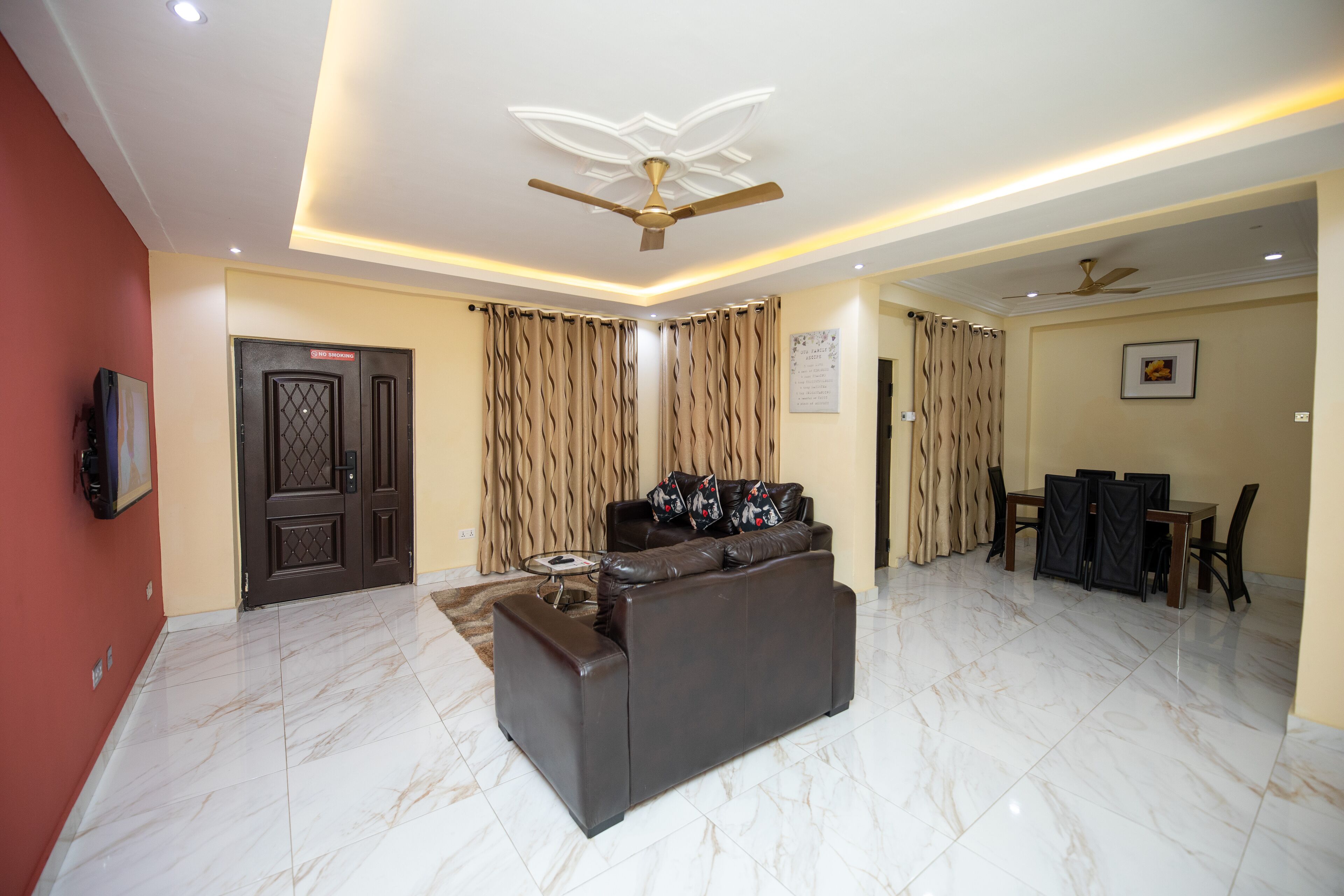A modern living room with a sofa, a dining table with chairs, a ceiling fan, and a TV mounted on the wall.