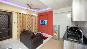 Una sala de estar moderna con un sofá, televisor, ventilador de techo y una zona de cocina con refrigerador y estufa.