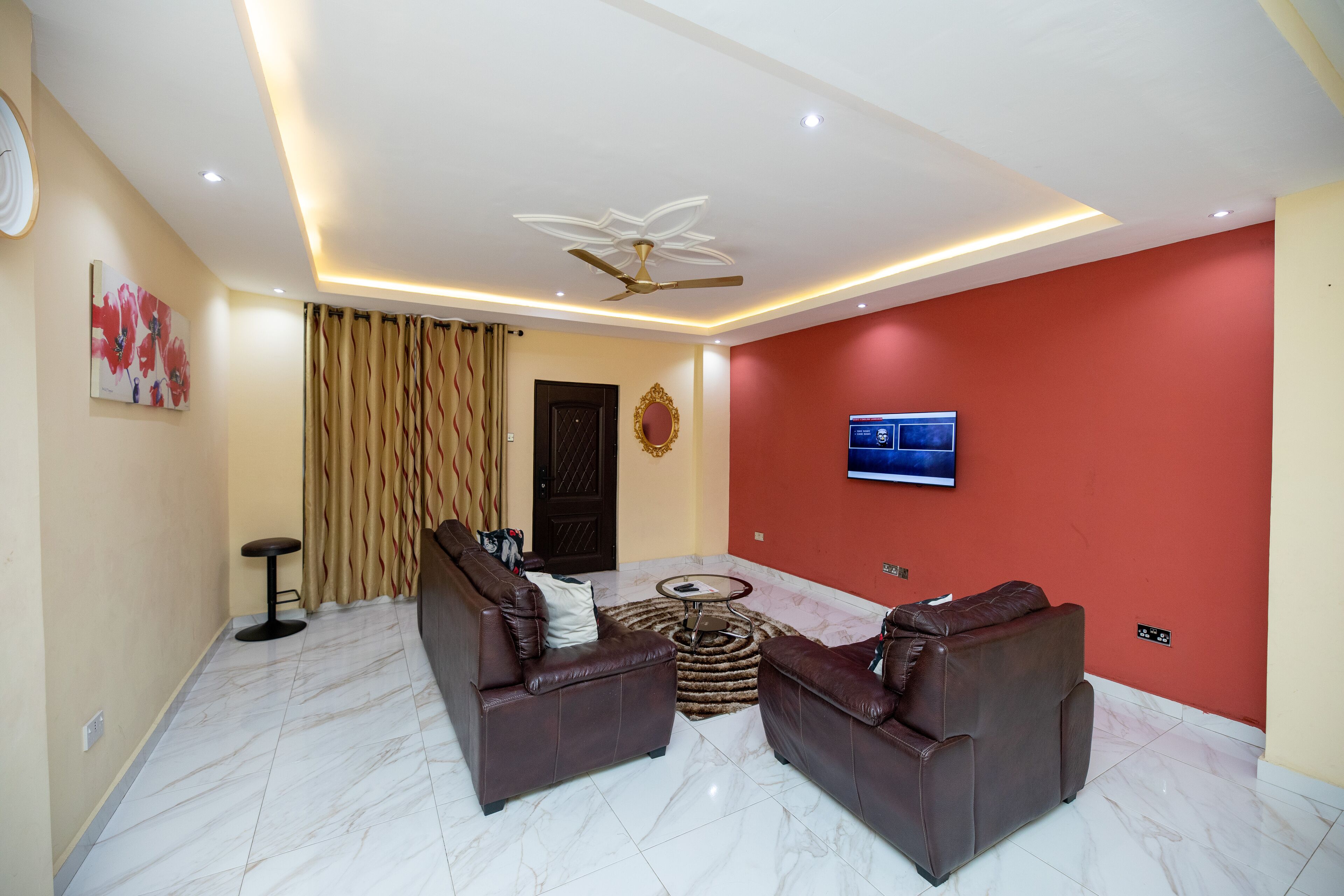 A modern living room with two leather sofas, a round coffee table, a ceiling fan, and a flat-screen TV mounted on the red wall.