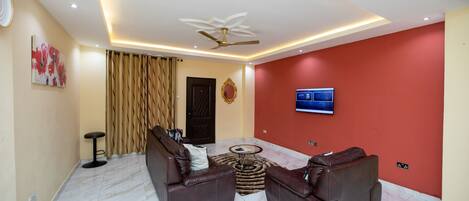 A modern living room with two leather sofas, a round coffee table, a ceiling fan, and a flat-screen TV mounted on the red wall.