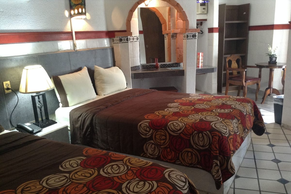A hotel room with two beds, a nightstand, a lamp, a mirror, and a small table with a chair.