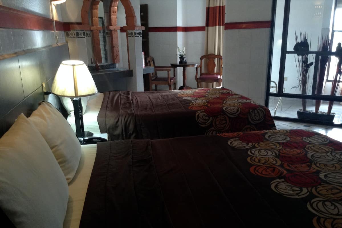 A hotel room with two beds, a nightstand, a lamp, a chair, and a window with a view of the outside.