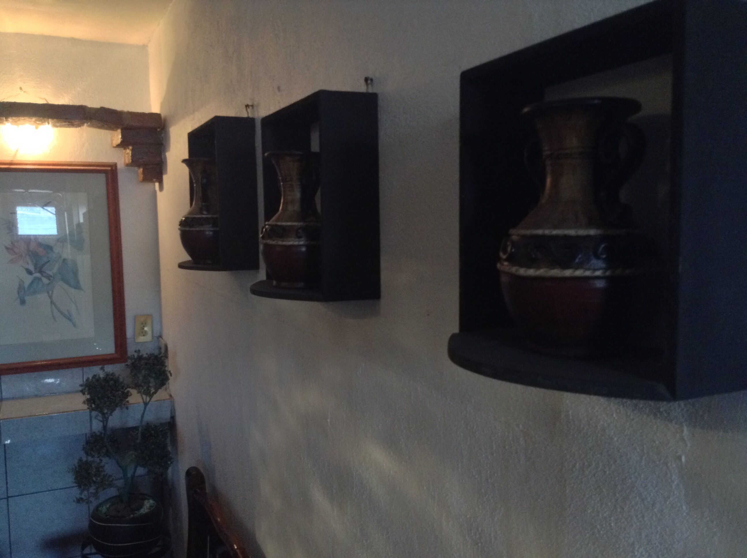 A wall with three dark wooden cabinets, each containing a large ceramic vase. There is a framed picture on the left and a potted plant below the cabinets.