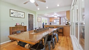 Un'ampia sala da pranzo con un lungo tavolo in legno e numerosi posti a sedere, una ventola a soffitto e un angolo cottura sullo sfondo.