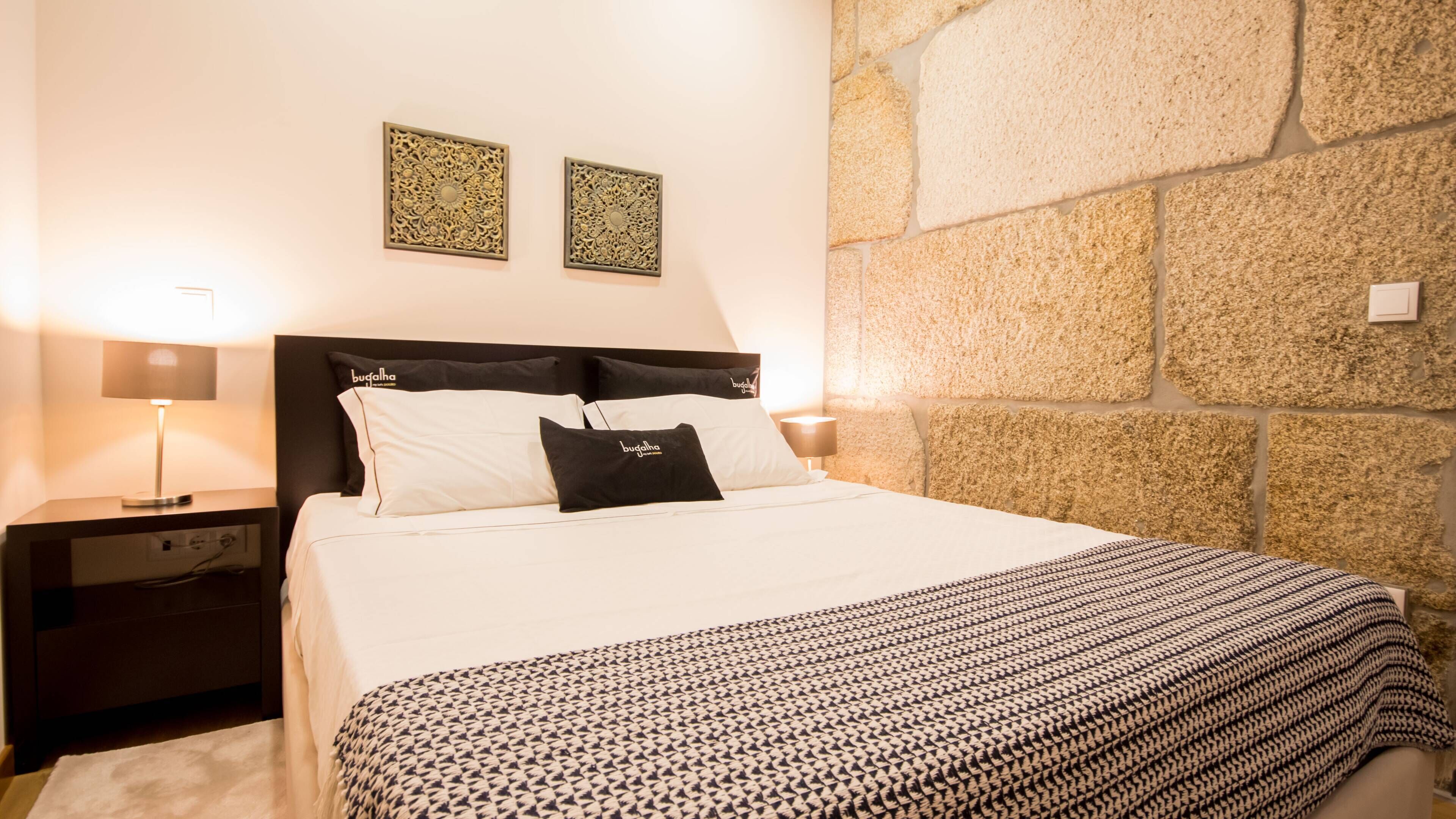 Una cama bien hecha con un edredón estampado, cabecera, dos paneles decorativos en la pared, mesita de noche con lámpara y pared de piedra.
