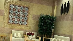 Um lobby com piso de azulejos bege, parede estampada, sofá de madeira, uma cadeira de madeira, uma mesa pequena com um vaso de flores vermelhas e uma grande planta verde.