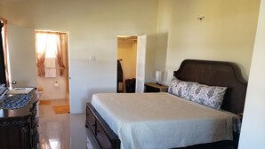 A bedroom with a bed, bedside table, and a dresser. There is a bathroom visible in the background.