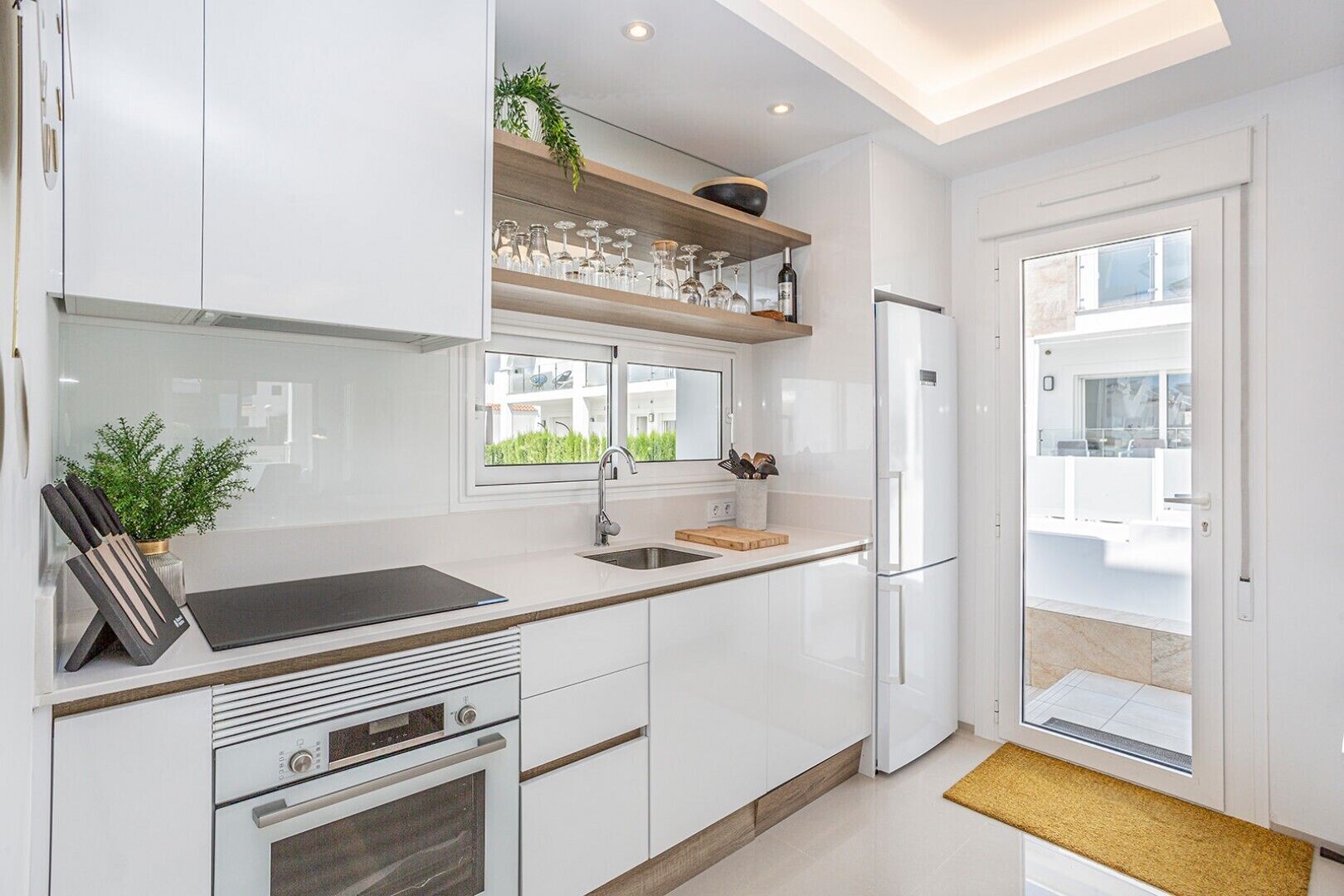 Een moderne keuken met witte kastjes, een inbouwoven, een spoelbak en een raam met uitzicht op een balkon.