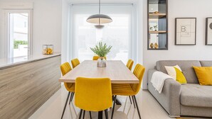 Moderni ruokailutila, jossa on puinen pöytä ja keltaiset tuolit, harmaa sohva ja suuri ikkuna, josta tulee runsaasti luonnonvaloa.