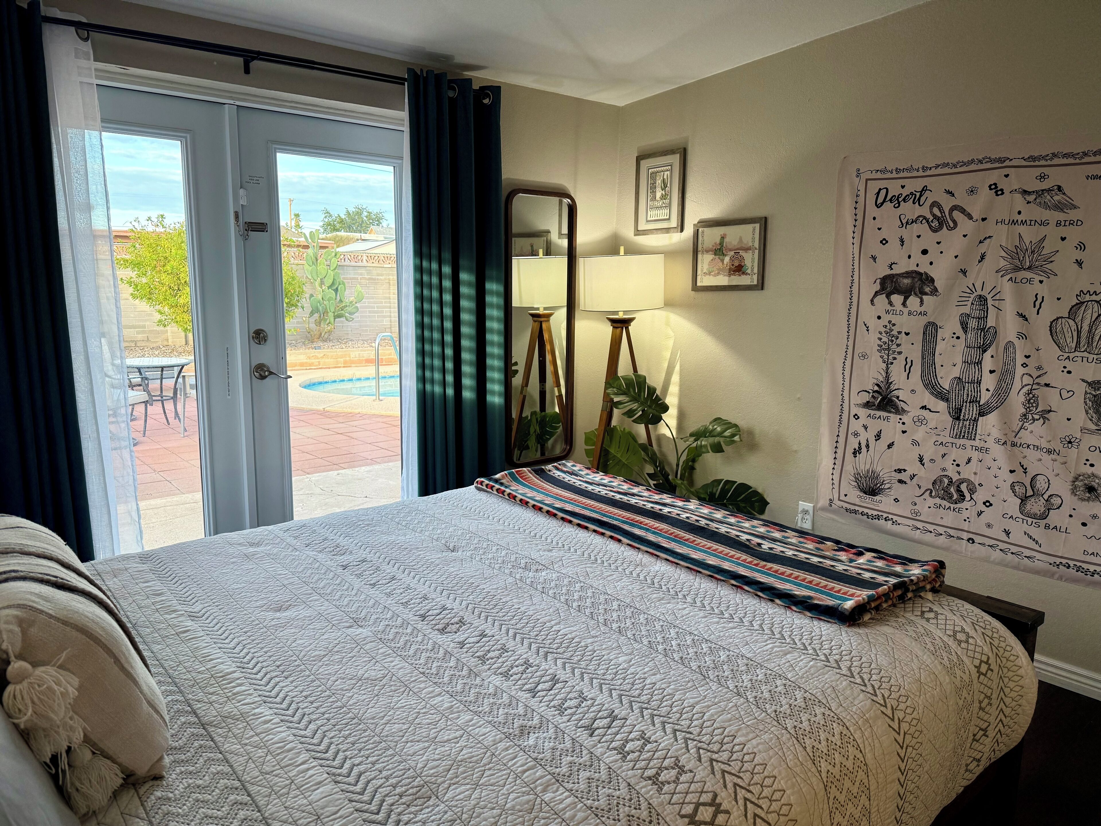 A bedroom with a bed, a large window with curtains, a mirror, a wall with a desert-themed poster, and a small table with a lamp.