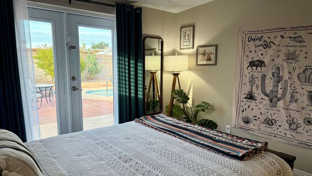 Um quarto com cama, uma janela grande com cortinas, um espelho, um mural com pôster de tema deserto e uma mesinha de cabeceira com abajur.