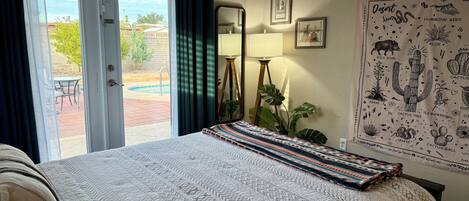 A bedroom with a bed, a large window with curtains, a mirror, a wall with a desert-themed poster, and a small table with a lamp.