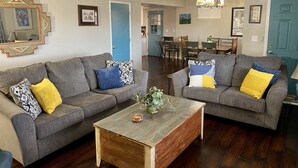Uma sala de estar com dois sofás cinzas, cada um com almofadas estampadas, uma mesa de centro de madeira e um ventilador de teto.