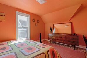 Une chambre avec un plafond en pente, un grand lit recouvert d’une courtepointe aux couleurs vives, une commode avec un miroir et une fenêtre donnant sur l’extérieur.