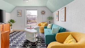 A cozy living room with a teal sofa, yellow armchair, and a white coffee table. A large window offers a view of the street. - 70's Vibe | Beach | Downtown | AC | Nightlife | (Traverse City)
